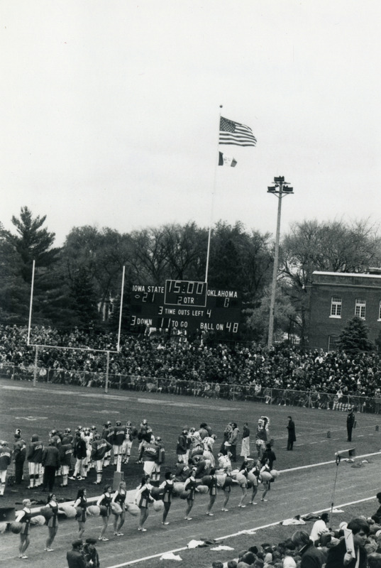 Cheerleaders are entertaining the crowd at the 1970 Homecoming Game. The scoreboard shows Iowa State leading over Oklahoma 21 to 0 in the second quarter.