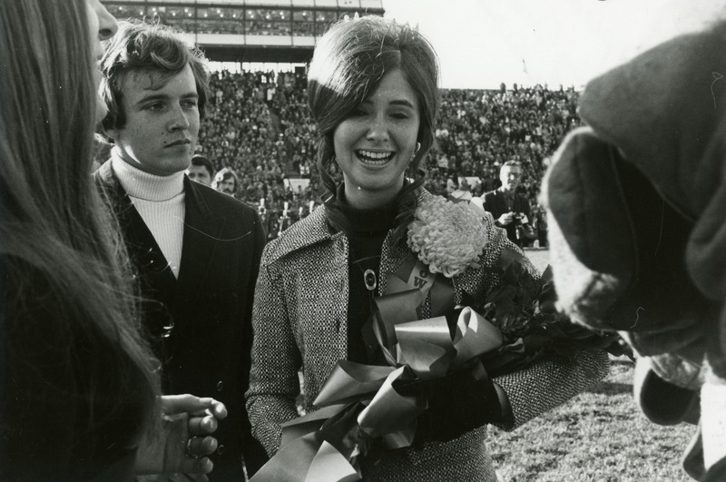 Diana Herbst, Homecoming Queen, is holding a bouquet of flowers as two students look on.