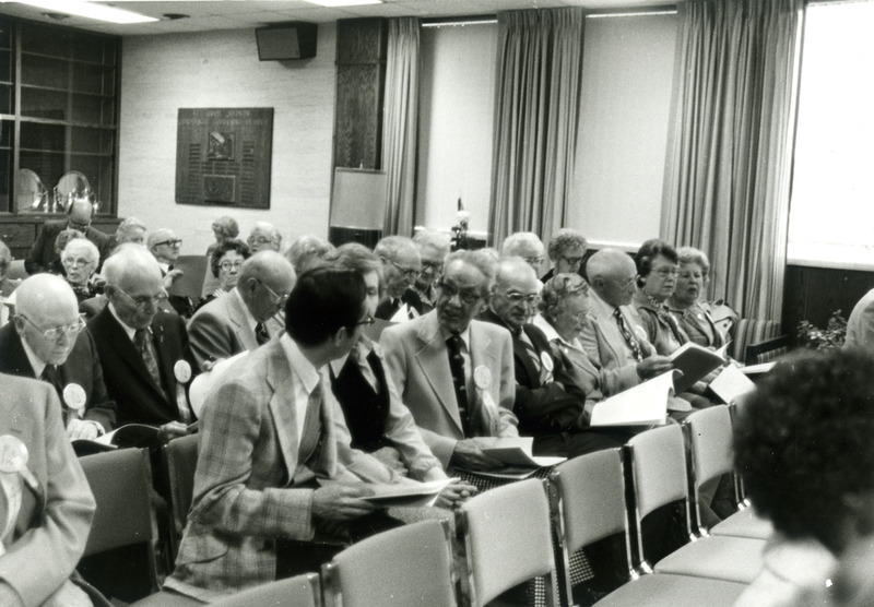 A group of alumni are seated and looking through booklets waiting for a program to begin.