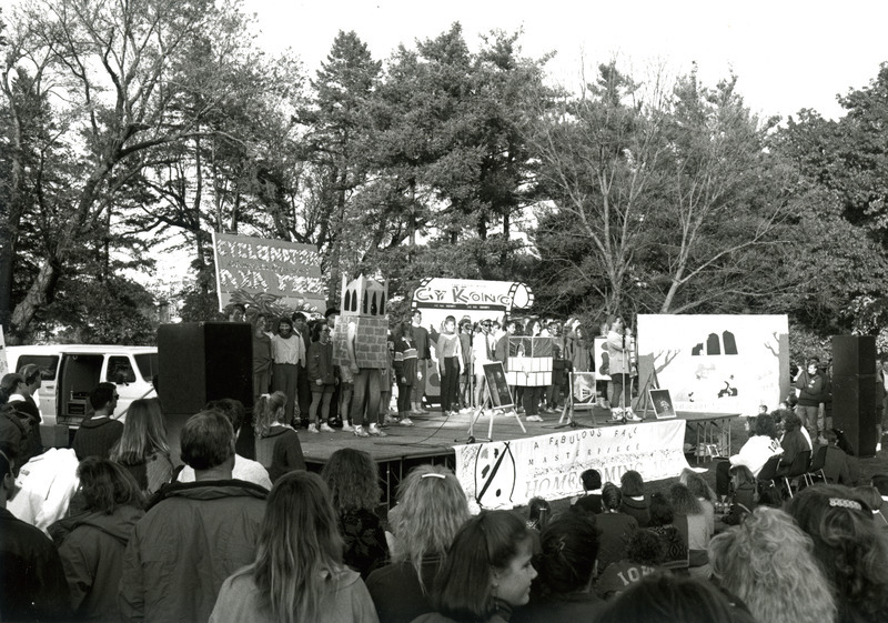 A student is speaking on central campus at the Student Organization Pep Council rally during homecoming. Some students on the stage have painted faces and are wearing cardboard costumes. The sign at the bottom of the stage reads, "A Fabulous Fall Masterpiece.".