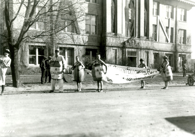 Five students dressed in wooden barrels are parading in front of the Dairy Building carrying banners and signs. One sign reads "A cyclone hit me." Two large letters, "A" and "N" appear on the second level of the Dairy Building.