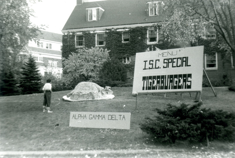 A student is looking at the Alpha Gamma Delta, Rho Chapter, lawn display showing a tiger laying between two sandwich buns. A sign on the lawn reads "Menu I.S.C. Special Tigerburgers.".