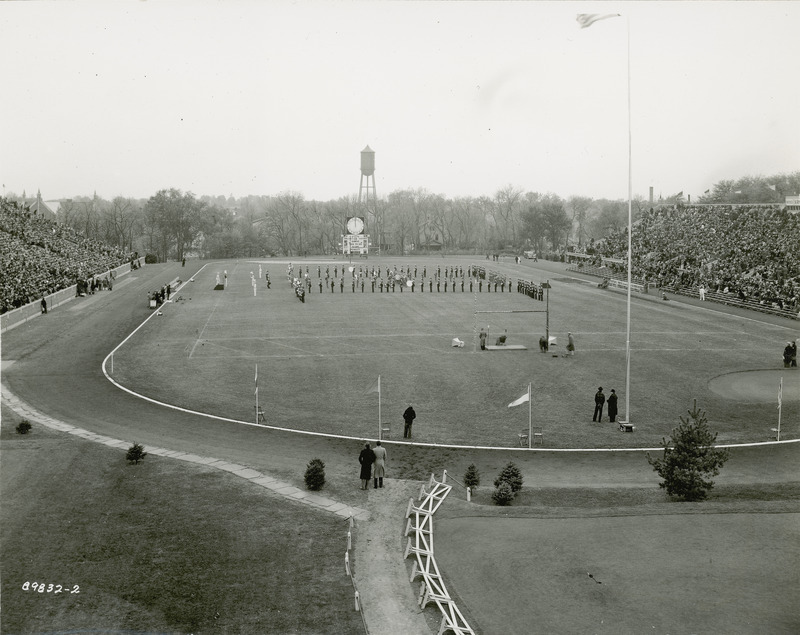 The ISU Marching Band is performing at the 1938 Homecoming football game at Clyde Williams Field. The Marston Water Tower is in the distance behind the scoreboard.