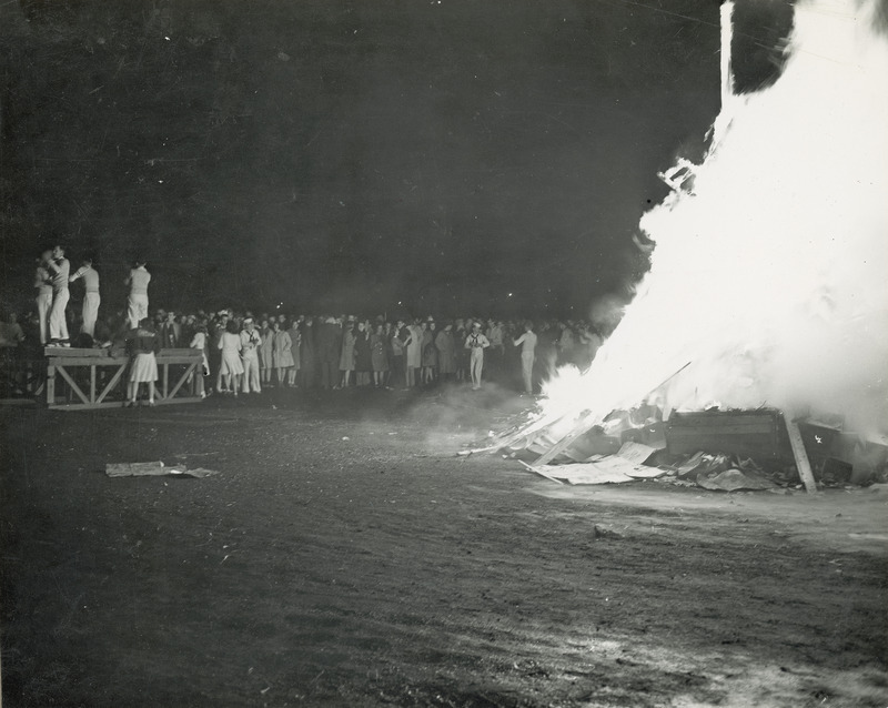 Students are gathered at the Homecoming bonfire. Some students are wearing sailor uniforms and others are standing on a stage.