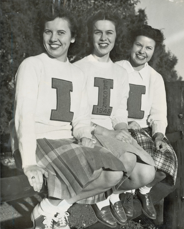 Betha Giffei, Pep Queen (center); Betty Lou Anderson (left) and Lucille Saunders (right) are sitting on a wooden fence. The women are wearing letter sweaters and Giffei has the words "Pep Queen 1945" sewn onto the "I" on her sweater.