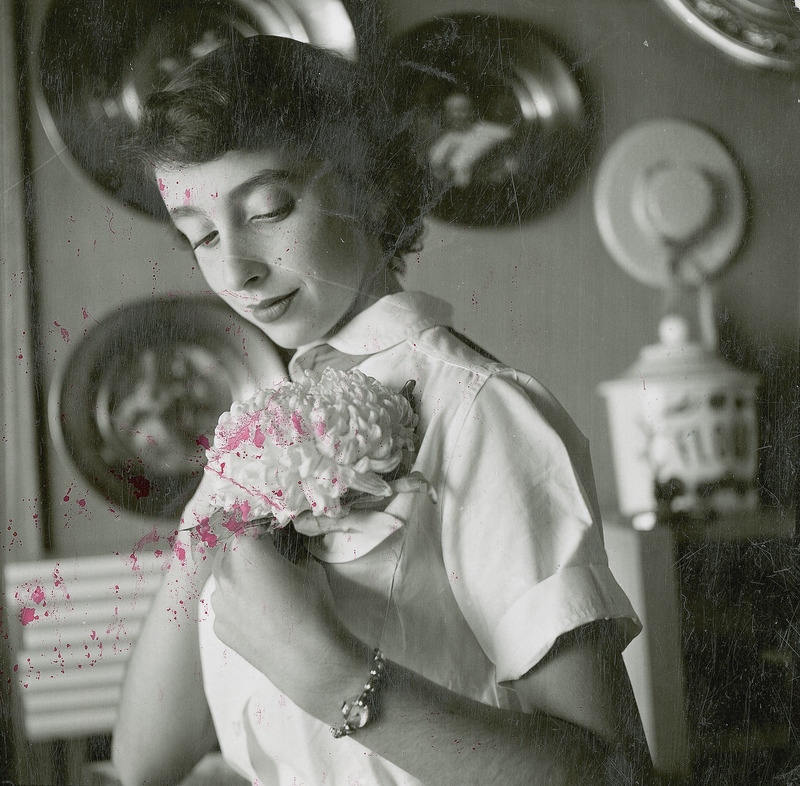 A girl is attaching a mum corsage to her blouse at Homecoming. Round picture frames and a woman's bonnet are hanging on the wall behind her.