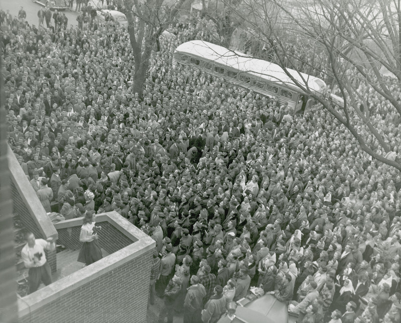 Two cheerleaders are leading a large crowd in a cheer at the Homecoming Pep Rally outside the Memorial Union. Football players are making their way through the crowd to a bus parked at the back of the crowd.