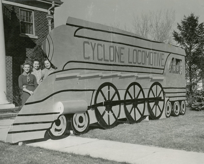 Three girls are posing with a lawn display of a train labeled "Cyclone Locomotive.".