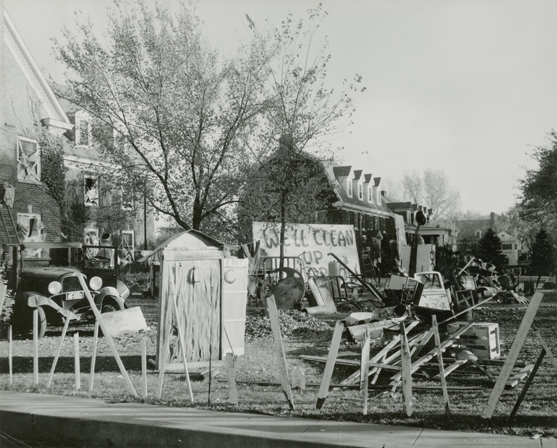 This Pi Phi House lawn display depicts a junk yard with a sign that reads, "We'll clean up tomorrow." An outhouse, old vehicle, and miscellaneous equipment parts are scattered on the lawn.