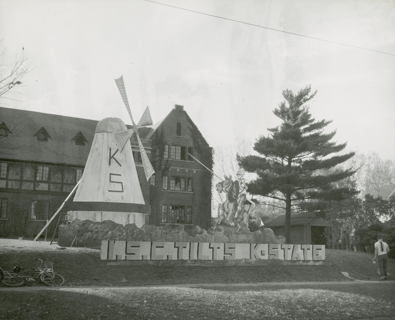 A large windmill with KS written on the front and an ISC sword-carrying knight on horseback are depicted in this Sigma Chi, Beta Omicron Chapter's lawn display. A sign across the front of the display reads, "I.S.C. tilts K-State." A student is standing on the right as a group gathers in front of the garage to view the display.