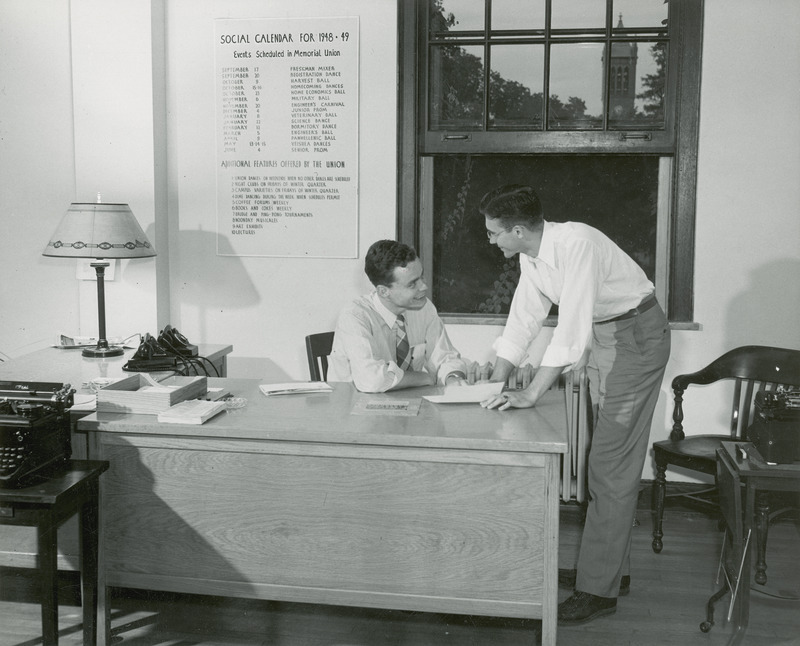 Two men are having a conversation in the Homecoming Office in the Memorial Union. The campanile can be seen through the open window behind them. The social calendar for 1948-49 is displayed on the wall.