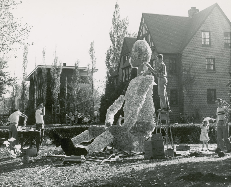 Students are working on a lawn display at the Alpha Chi Pho house. Two students are standing on ladders in the center of the photograph.