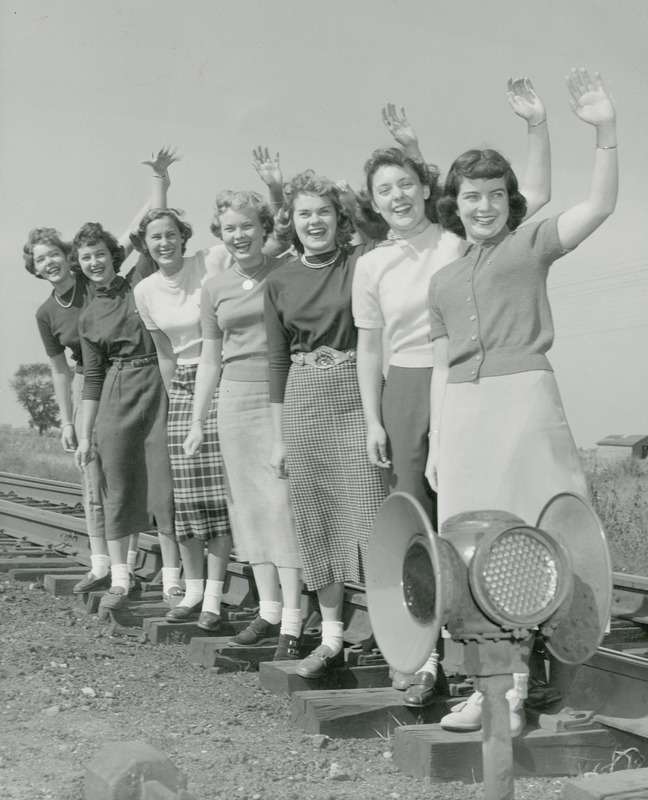 The 1950 homecoming queen candidates, waving from the railroad tracks, are (left to right) Marlene Sidney, Davenport; Janet Green, Sac City; Pat Binder, Barrington, IL; Ethel Pearce (Attendant), Mesa, AZ; Betty Skyberg (Attendant), Canton, SD; Claire Schubert, Maywood, IL; Rita Dunn (Queen), Sioux City.