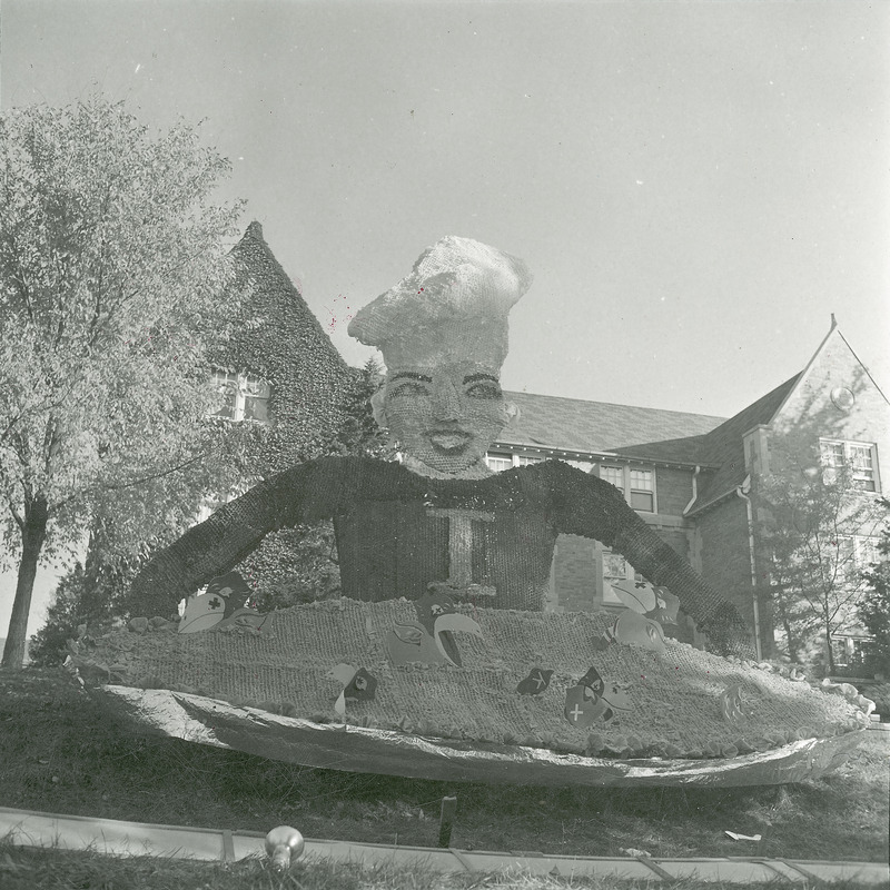 A tall woman figure is wearing a chef's hat and is holding a pie in this Alpha Delta Pi lawn display. Several University of Kansas Jayhawk figures are scattered throughout the pie.