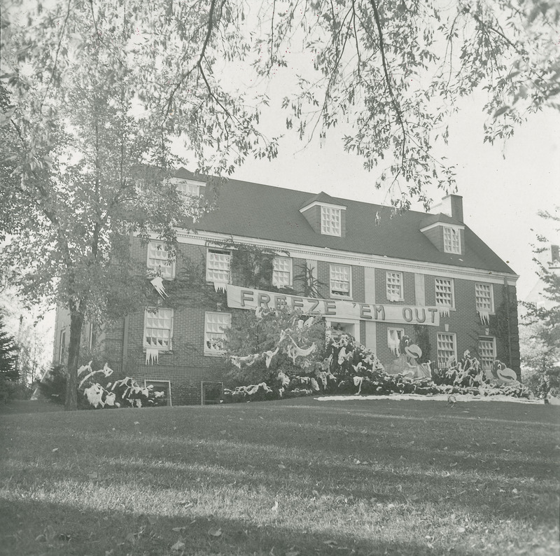 The windows and bushes at the Alpha Gamma Delta, Rho Chapter house are decorated to depict a winter scene. Two Kansas Jayhawk figures are sitting among the frozen landscape. A banner reads, "Freeze 'em out.".