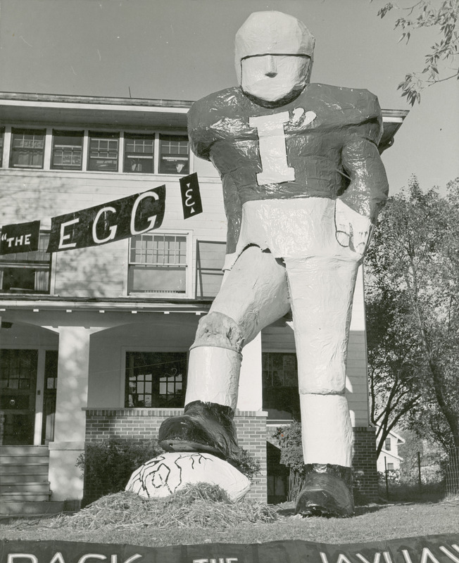 A sign reading "The egg &" points to the "I" on the jersey of a large football player standing on the lawn of the Delta Tau Delta, Gamma Pi Chapter house. The player is stepping on an egg. The banner "Crack the Jayhawks" can be partially seen at the base of the photograph.