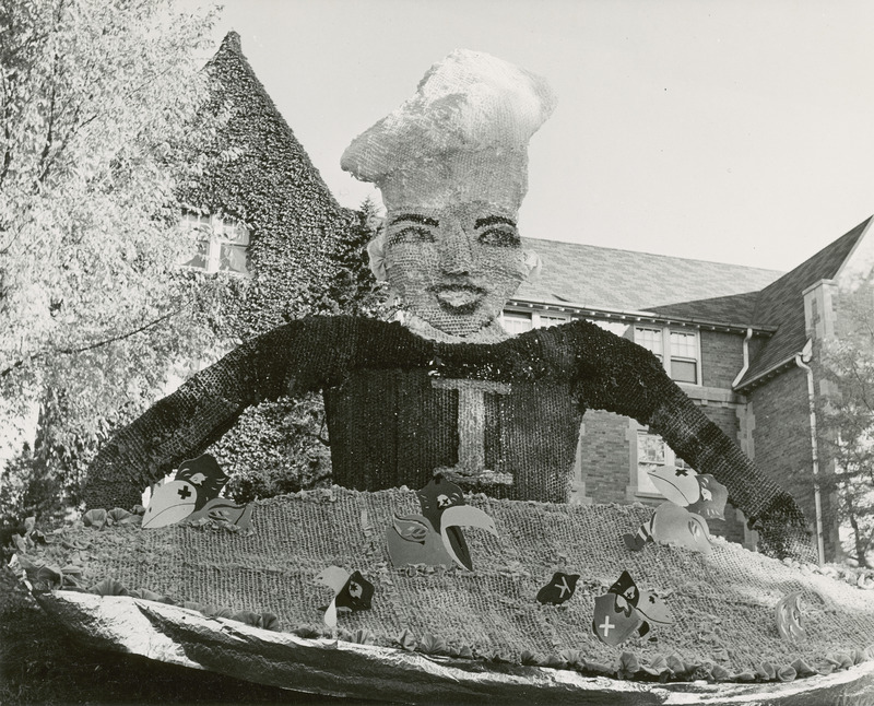 A tall woman figure is wearing a chef's hat and is holding a pie in this Alpha Delta Pi lawn display. Several University of Kansas Jayhawk figures are scattered throughout the pie.