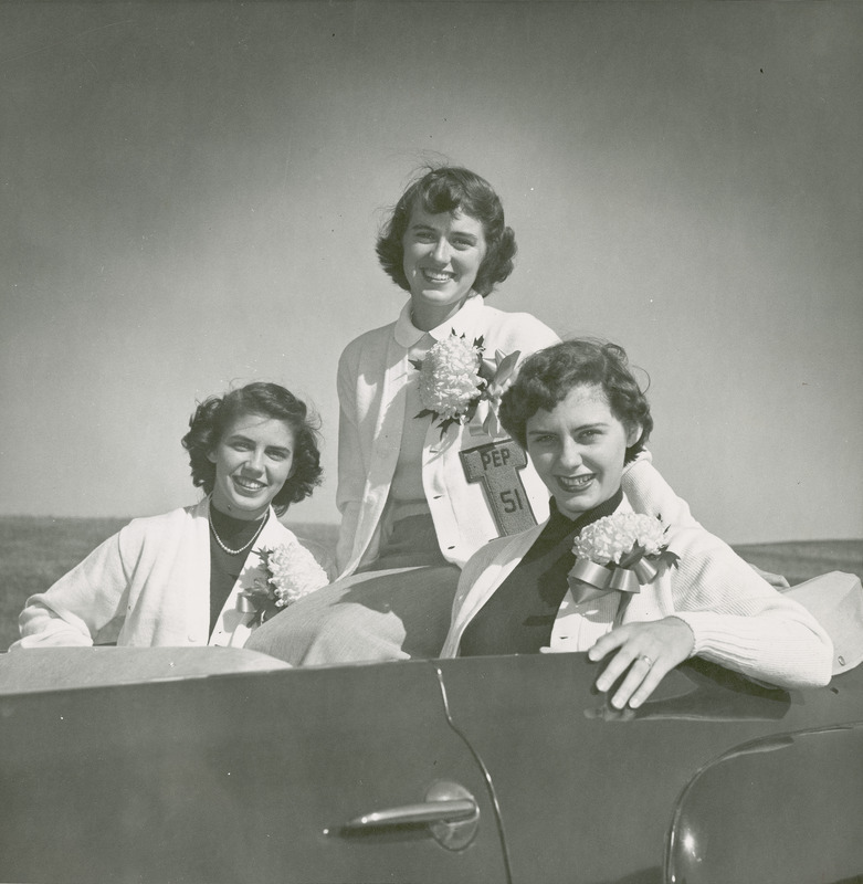 The 1951 Homecoming Queen and attendants, wearing mum corsages, are sitting in a convertible. They are from left to right : Ann Bradley (Attendant); Doris Blair (Queen); and Bonnie Helfrich (Attendant).