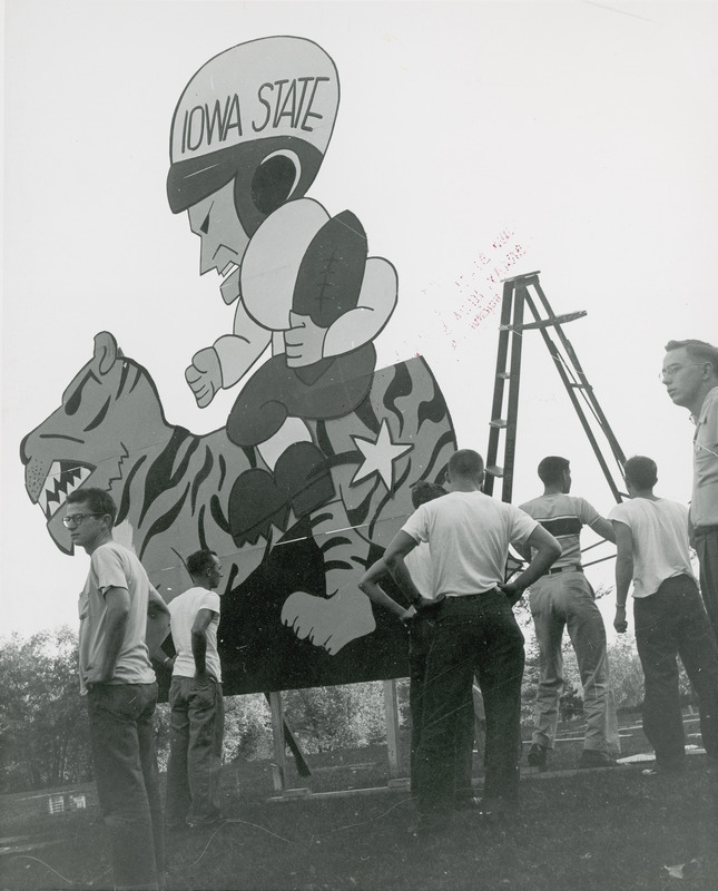 Several male students are gathered around a Friley Hall lawn display of an Iowa State football player taming a Kansas State wildcat in a Wildcat Rodeo.