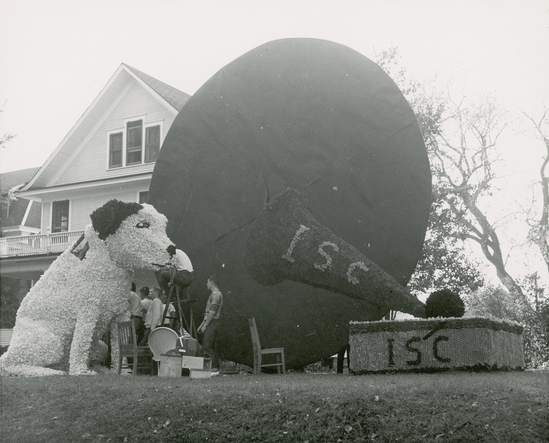 Cy the Dog sits attentively listening to a vintage phonograph player in this lawn display. A large dark disc is located behind the dog and phonograph. Several people are working on this Lambda Chi Alpha lawn display.