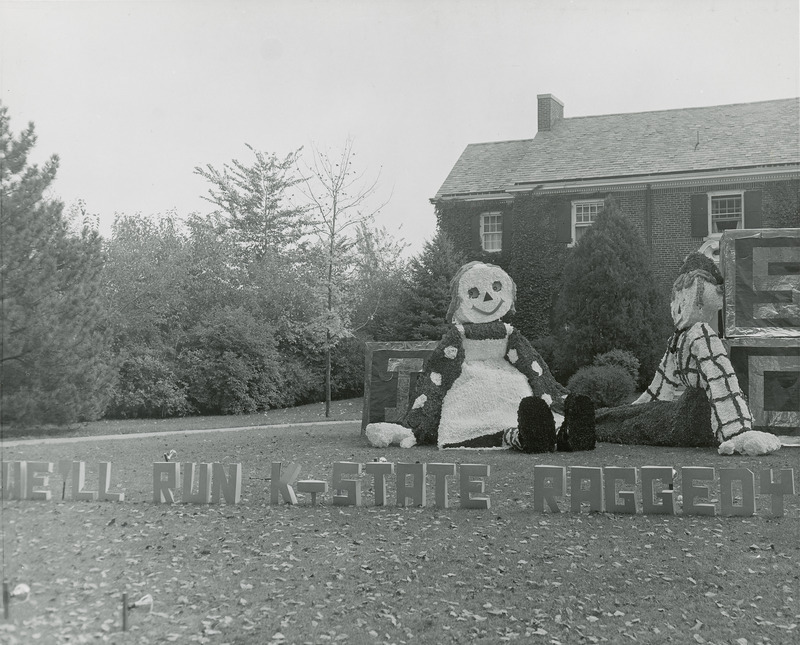 The Kappa Kappa Gamma, Delta Omicron Chapter lawn display features Raggedy Ann and Andy characters sitting on the lawn among blocks labeled I, S. C. Letters placed on the ground spell out, "We'll run K-State Raggedy.".