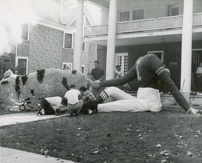 Phi Kappa Phi, Alpha Omicron Chapter fraternity members are working on Homecoming decorations. A headless football player holding the tail of a cat is part of the lawn display.