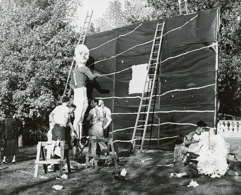 Students are working on the Sigma Phi Epsilon, Iowa Beta Chapter lawn display. Some ladders are leaning against a large banner with white horizontal lines. The figure of an ISC football player, facing the banner, is turning his head to the side.