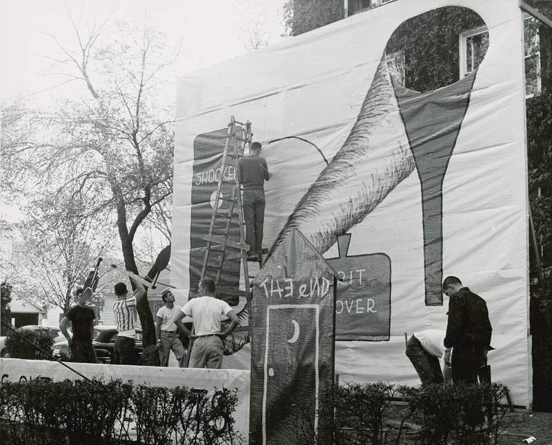 Students are working on the Phi Delta Theta, Iowa Gamma Chapter lawn display. A student is on a ladder drawing on the banner. A cyclone, funnel and outhouse are features included in the display.