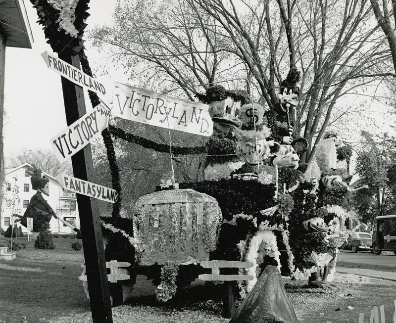 In this homecoming lawn display, Disney characters including Scrooge McDuck and Mickey Mouse ride with Cy in a jalopy heading for a signpost pointing to Victoryland, October 1961.