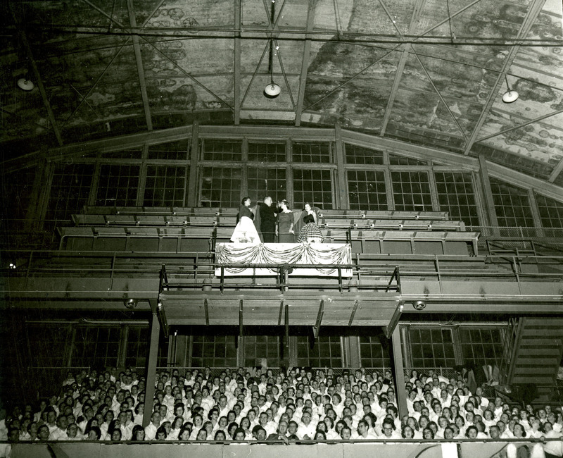 Cathy Olson is crowned 1957 Homecoming Queen by Chuck Schwab at the Pep Barbeque in the Armory. The attendants, Linda Sloan and Jo MacDonald look on. The Iowa State Singers pay tribute by singing "Friendly Persuasion.".