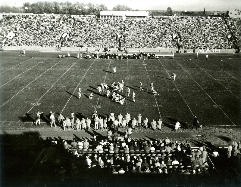 Football players on the field make a play during the Homecoming football game, viewed looking east from high in the stands, 1959.