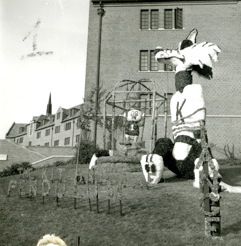 Lange House lawn display by Friley Hall, labeled The Kit and the Pendulum, features the Kansas State University Wildcat tied up by Cy as Tweety Bird, swinging in a bird cage with a saw poised over the cat's tail, October 1961.