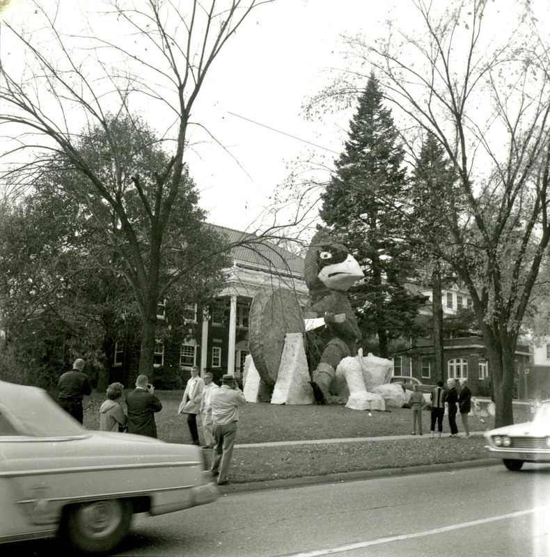 Beta Theta Pi, Tau Sigma Chapter, lawn display of Let's Skin 'Em Again, with Cy sharpening a blade while sitting on the Kansas State University Wildcat, viewed from Lincoln Way, October 1961.