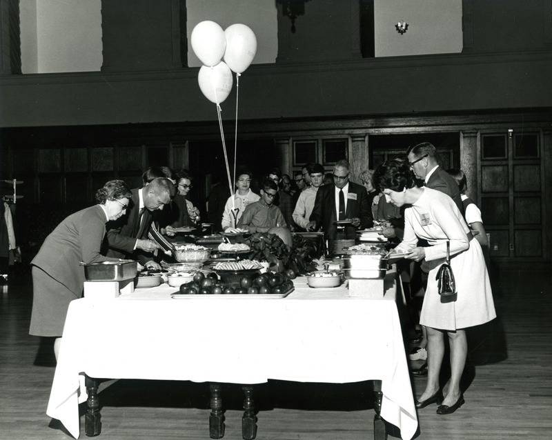 Alumni line up on both sides of a table for a homecoming buffet luncheon, 1966.