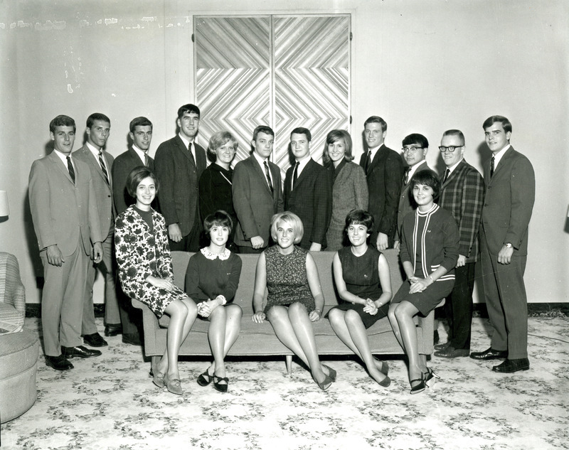 The seven women and ten men of the Homecoming Central Committee sit on or stand behind a sofa, 1966.