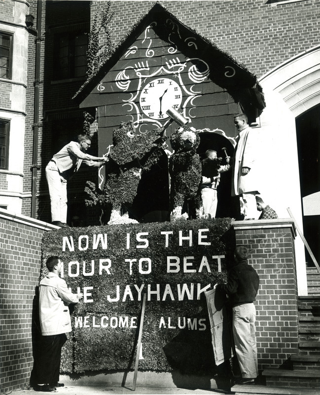 The Stevenson House Homecoming display of "Now is the Hour to Beat the Jayhawks" won first place in the Men's Residence Association, 1958. Students are shown working on the display, located in front of Friley Hall. It features a large cuckoo clock in which Cy strikes the Kansas University Jayhawk when both emerge from the clock.