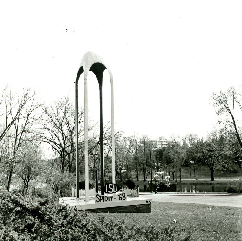 The second Homecoming monument built by Delta Chi and Alpha Gamma Delta features an open tower and signs saying ISU Spirit of '68 on all four sides. It overlooks Lake Laverne, on which a display shaped like a sailing ship can be seen in the background, 1968.