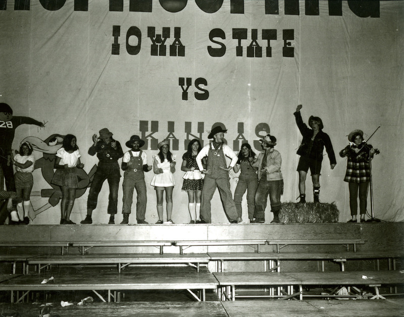 Twelve students dressed in country costumes clap and dance to a fiddler playing a country song, on the night before Homecoming, 1969.