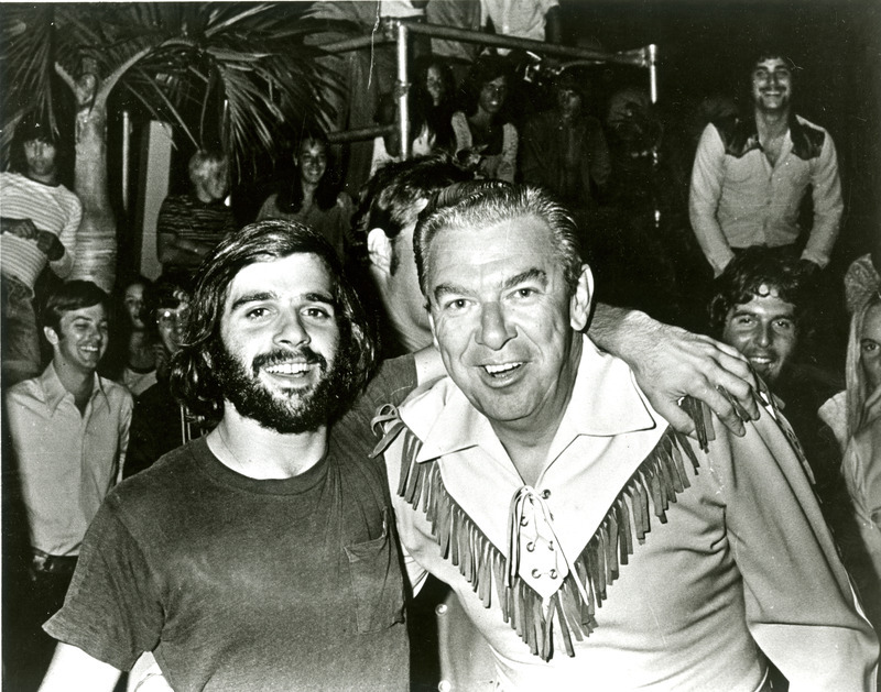 A bearded student puts his arm around Buffalo Bob, while other students look on happily in the background, at Homecoming, 1973.