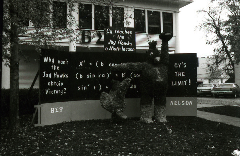 In the Beta Sigma Psi/Nelson House Homecoming lawn display, Cy holds a football away from a Kansas University Jayhawk while pointing to math formulas on a blackboard explaining "Why can't the Jay Hawks obtain Victory?", 1977.