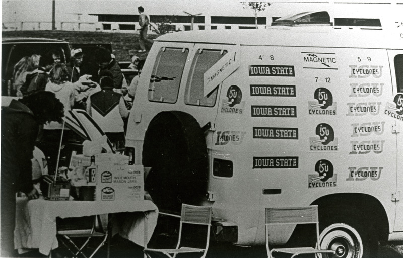 Cyclone fans gather around a table near a van decorated with Cyclone decals for Homecoming, 1980.