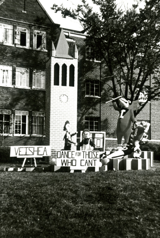 "Dance for Those Who Can't" on this Homecoming lawn display refers to the Muscular Dystrophy Dance Marathon that occurs close to Veishea. With a model of the campanile in the background, two dolls are shown dancing as well as two football players tackling each other, while Cy directs events with a baton.