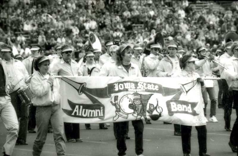 Three people carry a banner announcing the ISU Alumni Band, marching behind them on the football field at Homecoming, 1985.