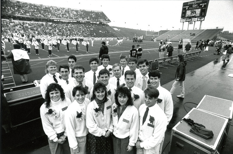 The Homecoming Central Committee for 1987 poses pregame by the football field with the ISU Marching Band performing in the background. They are left to right, 1st row: Julie Newman, Laura Butler, Jan Harnel, Heidi Hova, Tim Schumann; 2nd row: Mike Capp, Todd Appenzeller, Darren Hiov, Darrel Burt, Todd Allard; 3rd row: Marty Epy, Terry Helphrey, Dan Jones, Craig Jodan; 4th row: Mark Weikel, Jim Carter, John Bauerle, Steve Geadelmann.