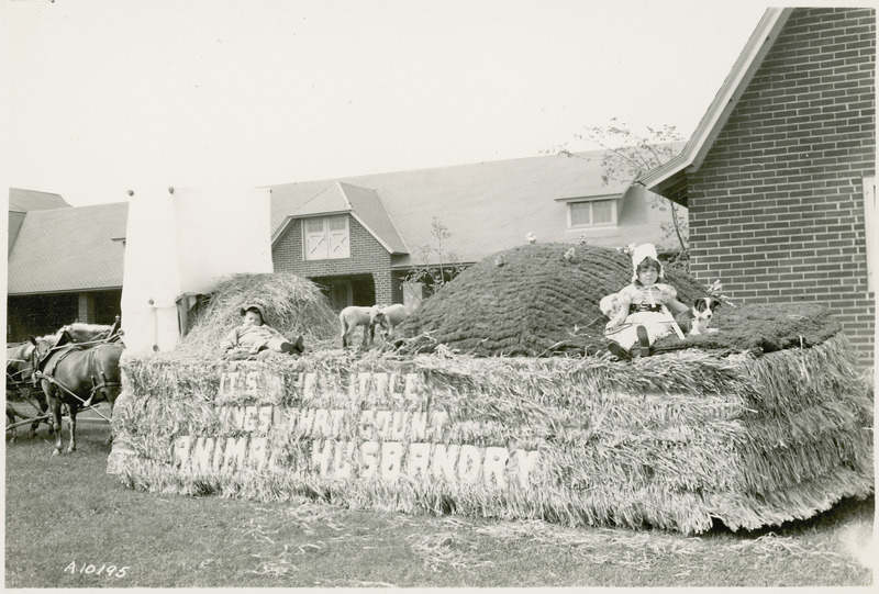 A 1938 VEISHEA parade float from the Department of Animal Husbandry. "It's the Little Things That Count." Two children rid atop this float with two lambs and a dog. Horse barns are in the background.