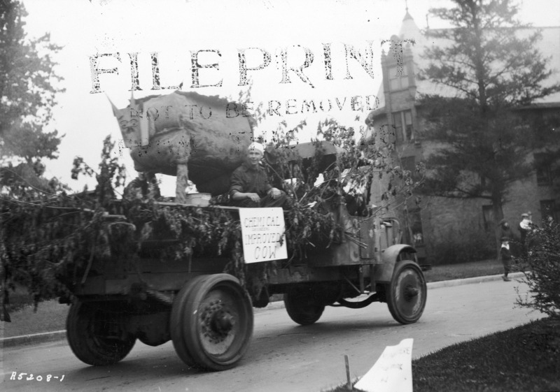 A 1923 VEISHEA float from the Department of Chemical Engineering. One student is sitting on the float which is covers with branches and topped with a large "cow." The sign reads: "Chemical Engineers Improved Cow.".