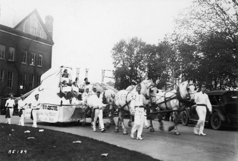 A 1923 VEISHEA horse-drawn float from the Department of Agriculture. Men in their dairying uniforms walk beside the float upon which six women are seated holding signs. Another woman is standing facing them. See photograph 1722-3-6 for another view of the same float.