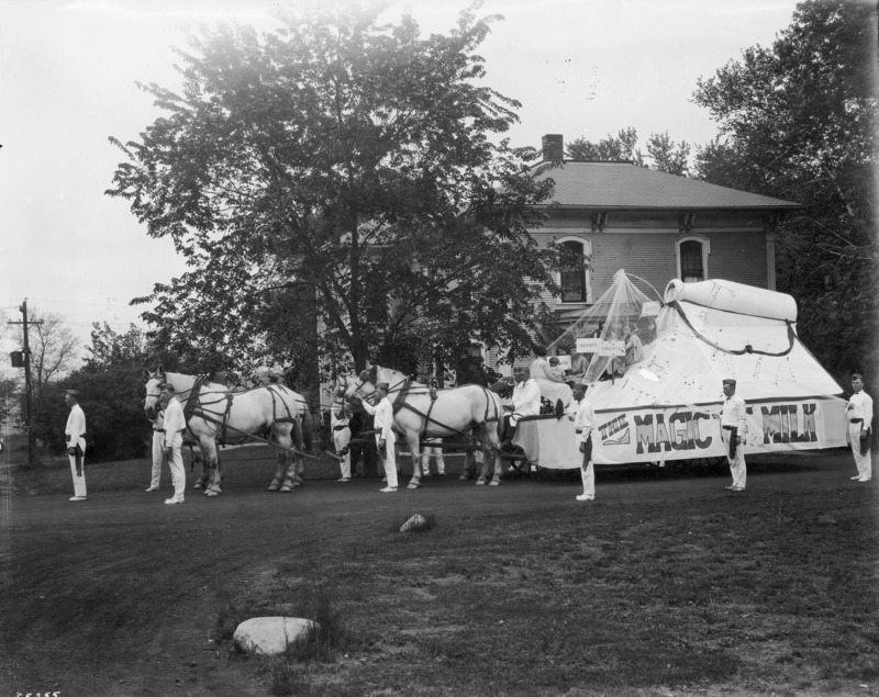 A 1923 VEISHEA horse-drawn float from the Department of Agriculture. "The Magic of Milk" is printed on the side of the float. Men in their dairying uniforms stand beside the float upon which seven women are seated. One woman has a child in her lap and the others are holding signs: "Growth," "Beauty," Laughter," and "Health." The float is topped with a giant milk bottle. See photograph 1722-3-5 for another view of the same float.