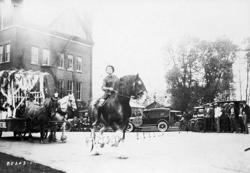 Man on draft horse wearing military uniform with tam and kilt. Period cars are visible in the background. See photograph 1722-4-1 for another views.