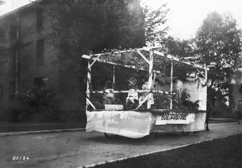 A VEISHEA Horticulture float. The float, built on a flat bed wagon, is a grape arbor in which are ferns and similar plant material. Three women are sitting behind the arbor. Margaret Hall is in the background.